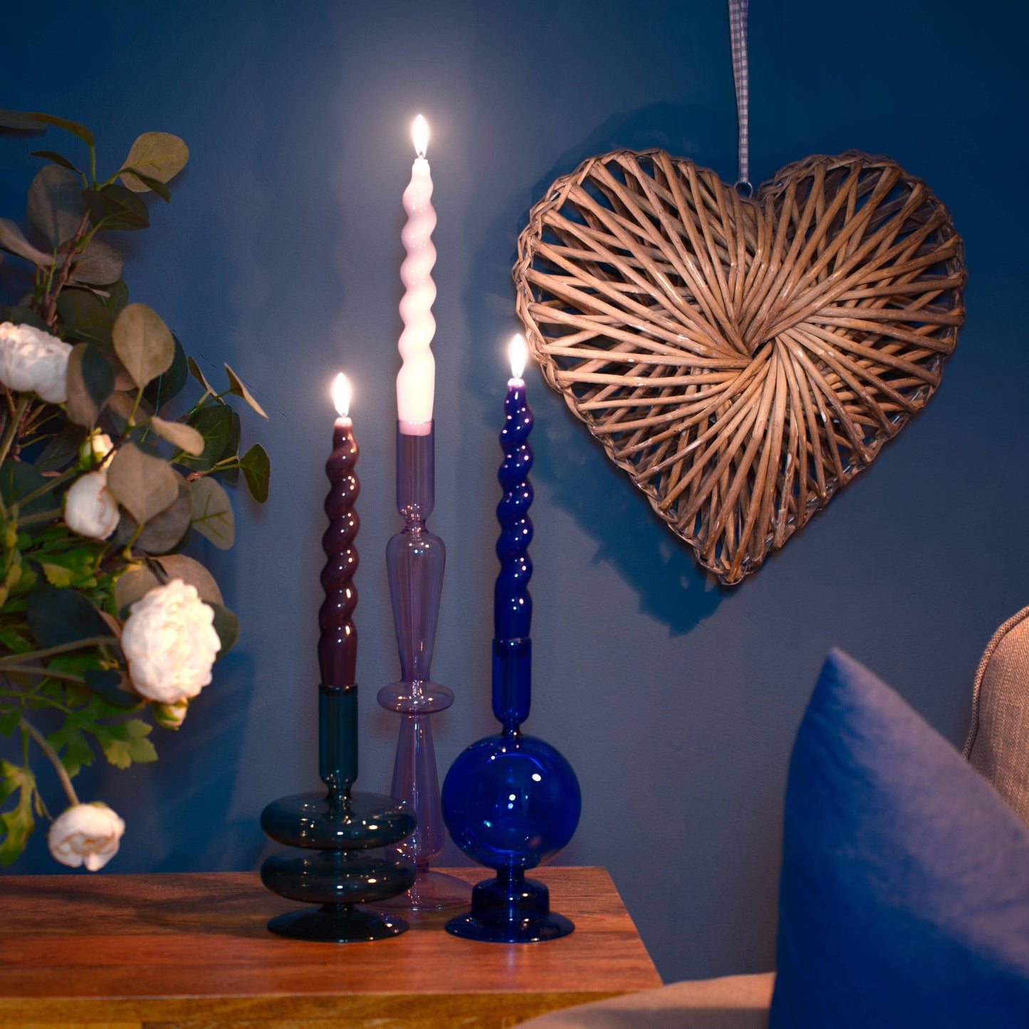 Decorative candles on a table with a heart-shaped wall hanging and a plant in the background.
