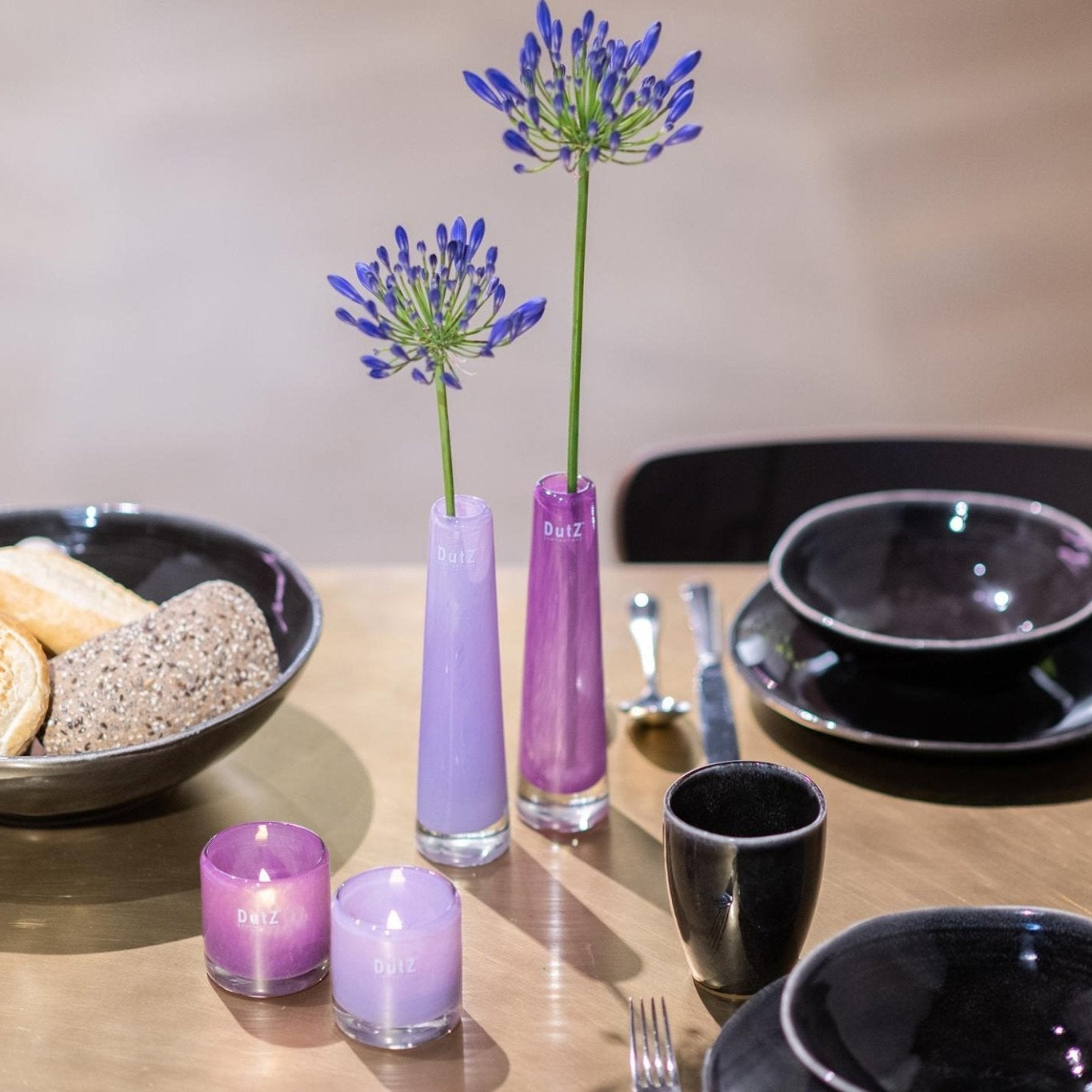 Dining table setting with black plates, purple candles, and a vase with flowers.