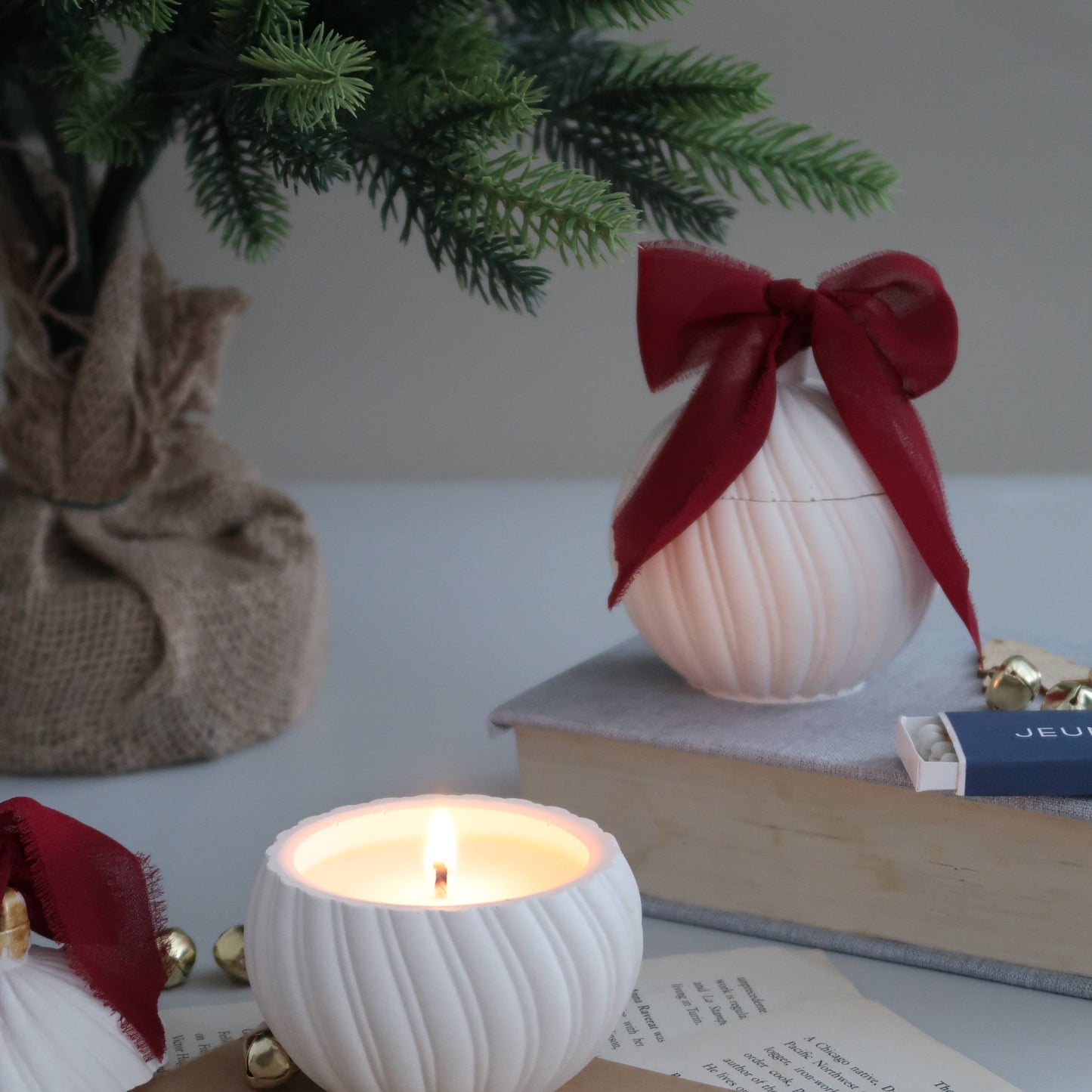 Candle in a textured white holder with a Christmas tree and decorative items in the background.