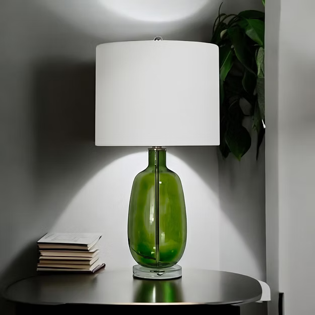 A green glass lamp with a white shade on a dark table, beside a plant and stacked books.