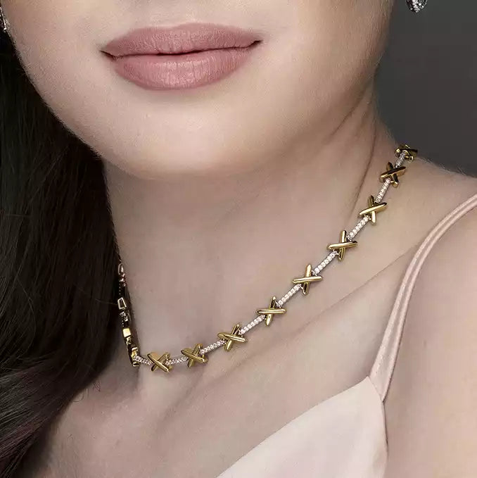 Woman wearing gold X necklace on light skin, with pink lips, dark hair, and pale pink top.