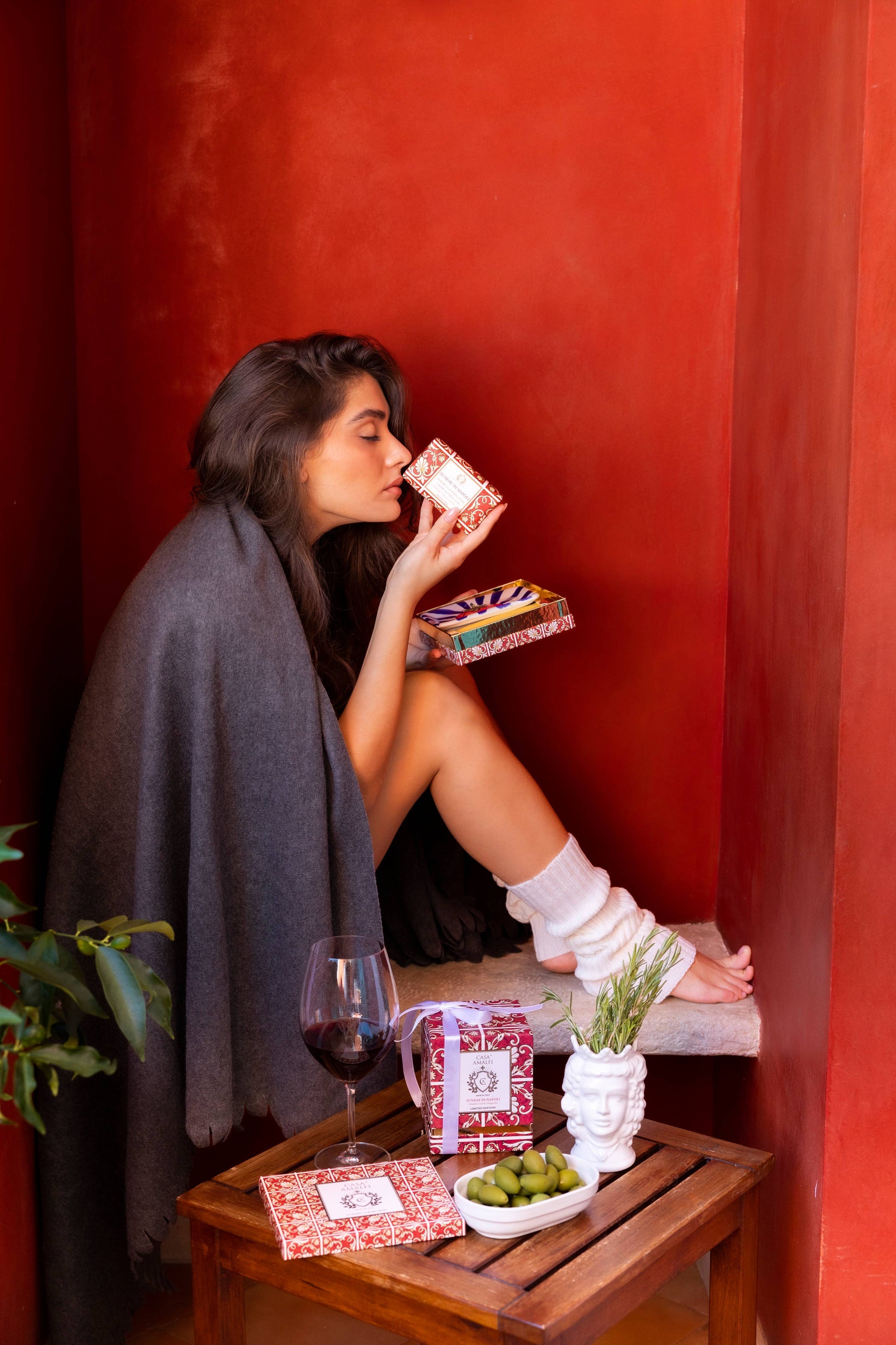 Woman sitting against a red wall with a glass of wine and snacks on a small table.
