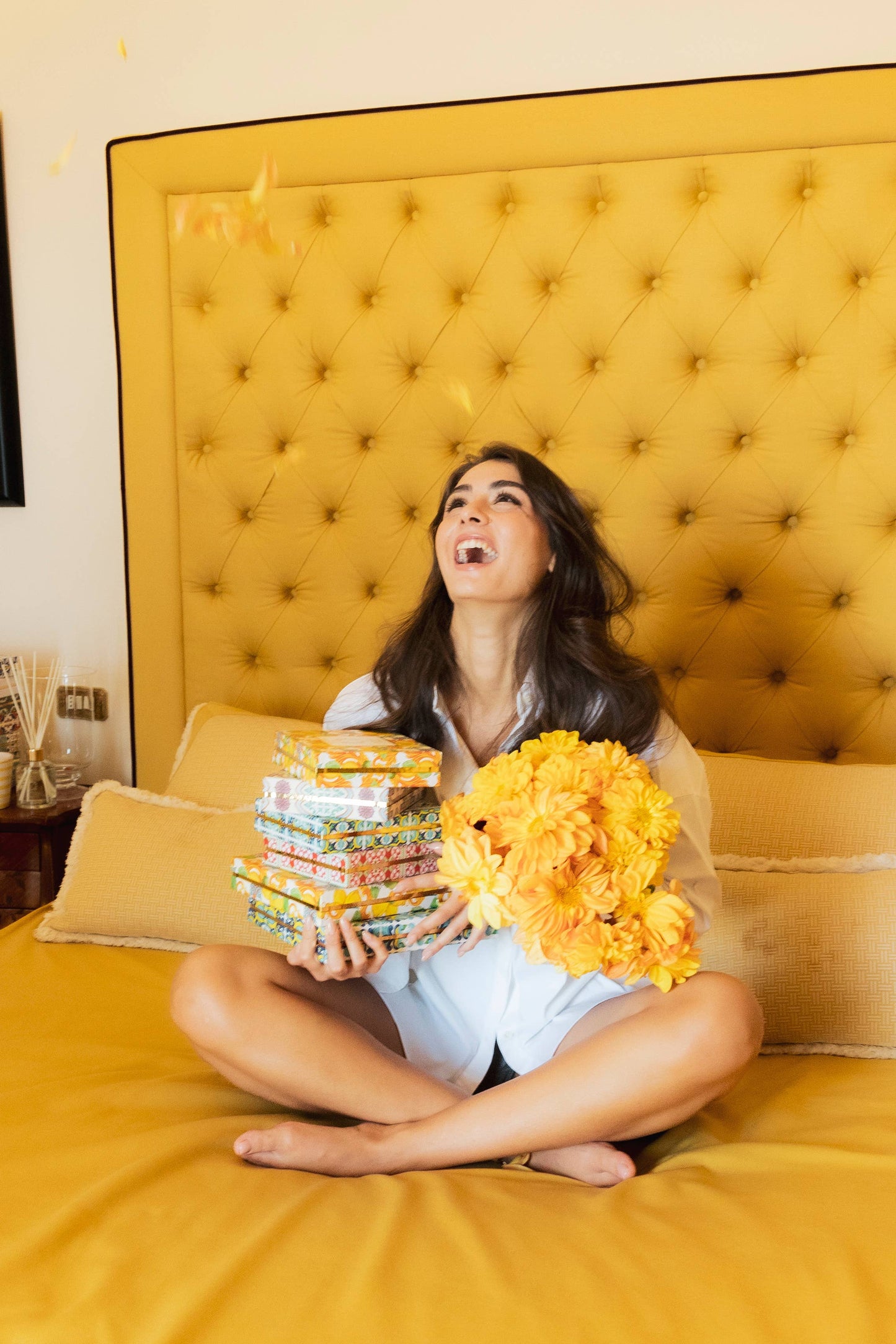 Woman sitting on a yellow bed holding books and flowers, laughing.