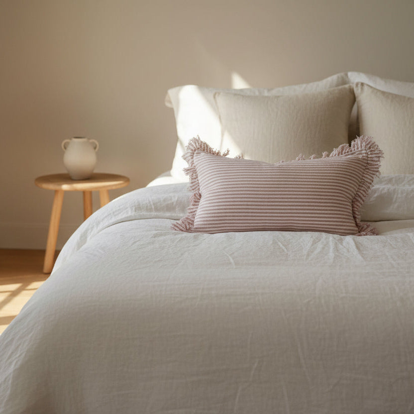 A rectangular 50x30cm pink striped linen cushion with a ruffled edge, styled in front of a larger white pillow on a bed.