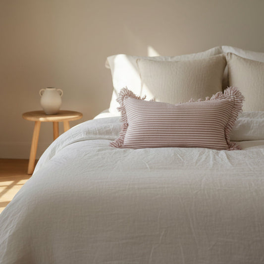A rectangular 50x30cm pink striped linen cushion with a ruffled edge, styled in front of a larger white pillow on a bed.