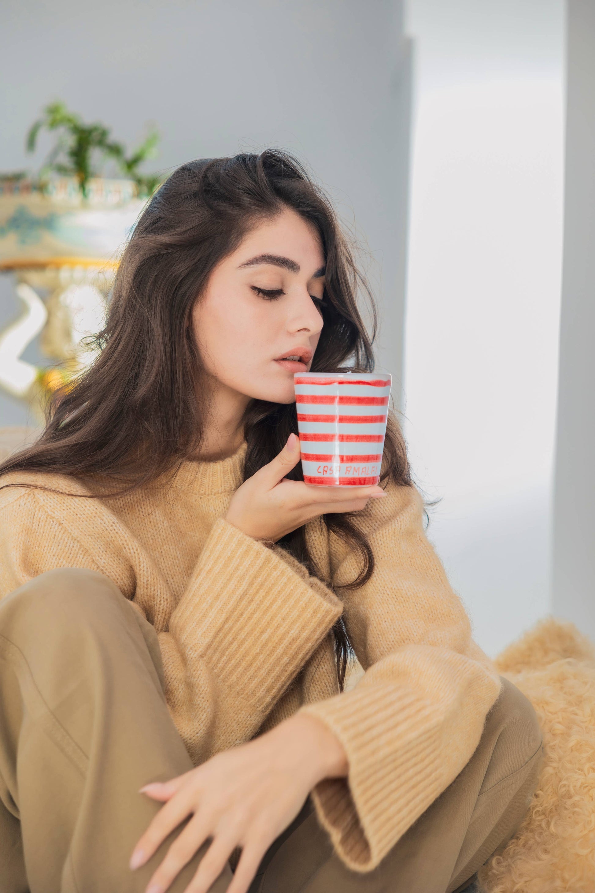 Woman in a beige sweater holding a red and white striped cup, sitting on a couch.