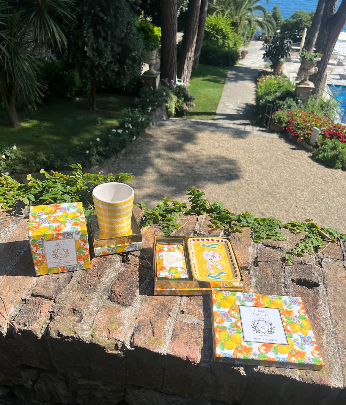 Decorative items including a cup, coasters, and a box on a stone ledge with a garden and pool in the background.
