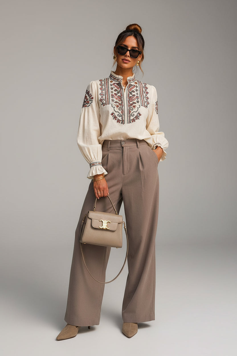 A woman stands outdoors against a beige stone building and black door, wearing a cream blouse with embroidered detailing at the chest, high-waisted taupe wide-leg trousers, and beige pointed-toe heels. She carries a taupe handbag.