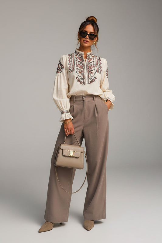 A woman stands outdoors against a beige stone building and black door, wearing a cream blouse with embroidered detailing at the chest, high-waisted taupe wide-leg trousers, and beige pointed-toe heels. She carries a taupe handbag.