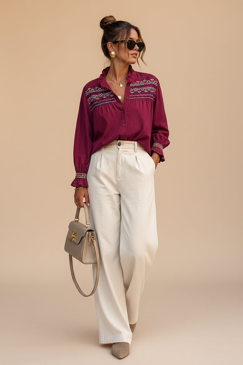A woman stands on a city sidewalk leaning against a beige stone building, wearing a burgundy long-sleeve blouse with colorful geometric embroidery across the chest and cuffs, paired with high-waisted off-white wide-leg trousers and beige pointed-toe heels. She carries a taupe handbag.