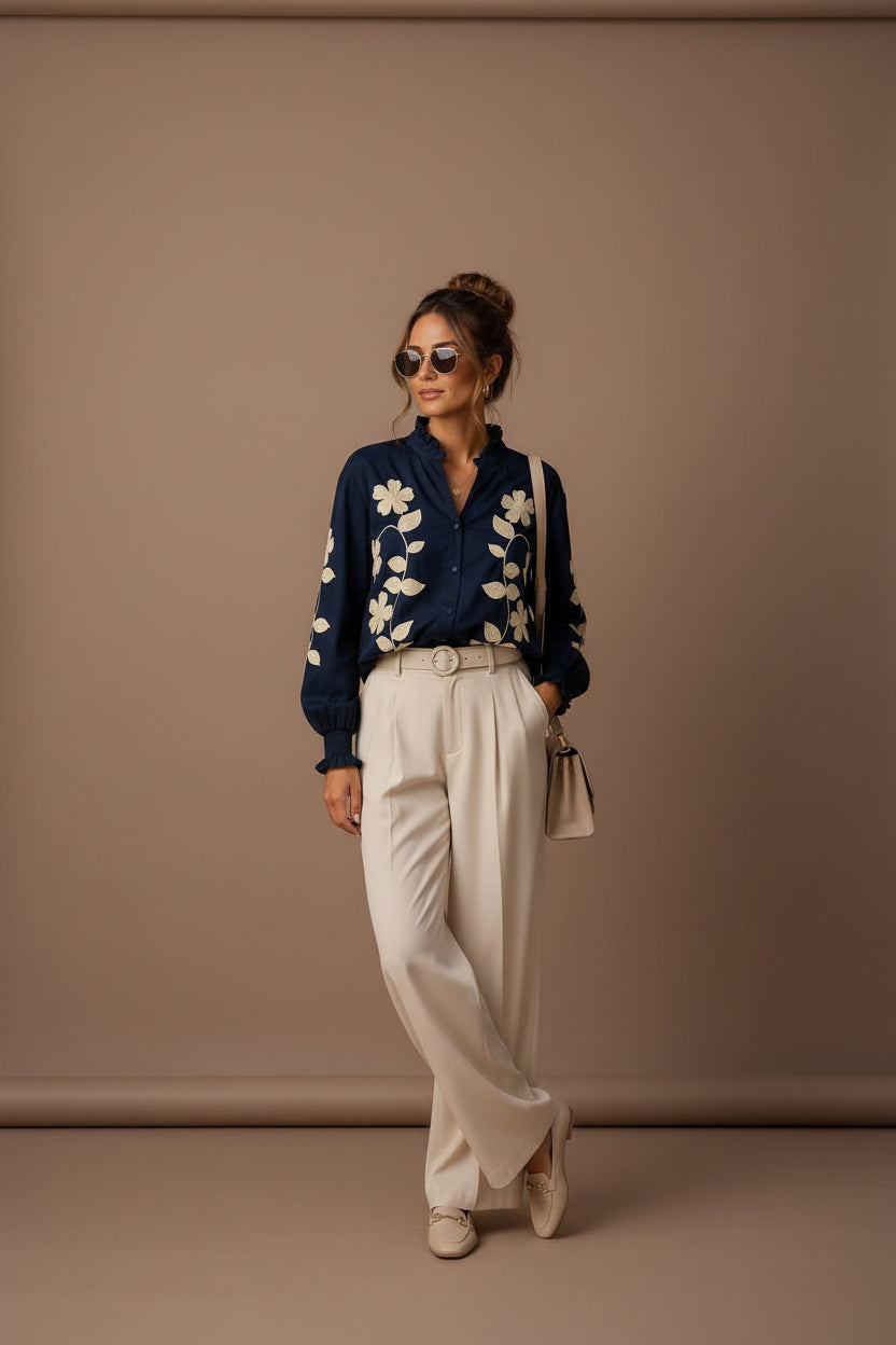 A woman stands on a city sidewalk wearing a navy long-sleeve button-up blouse with large cream floral embroidery, cream wide-leg trousers, beige loafers, and carrying a small beige shoulder bag.