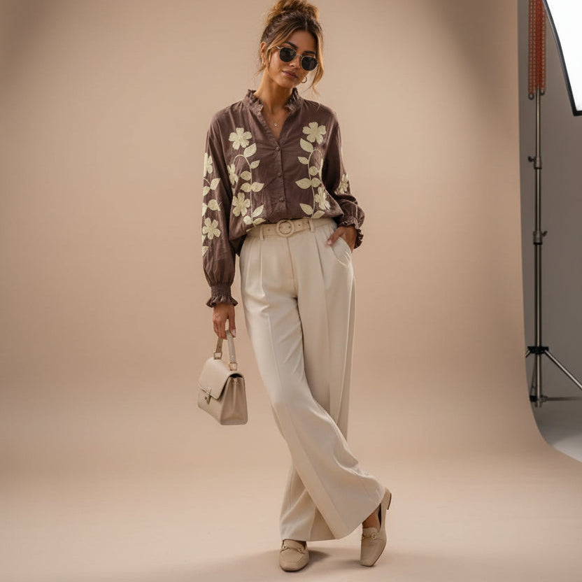 A woman stands on a city sidewalk wearing a brown long-sleeve button-up blouse with large cream floral embroidery, paired with cream wide-leg trousers and beige flats. She holds a small cream handbag and wears dark sunglasses, posing in front of a beige stone building and a black door.