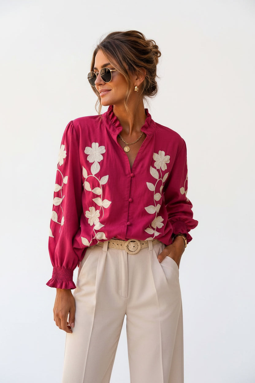 A woman wearing a vibrant magenta long-sleeve blouse with white floral embroidery, paired with cream-colored trousers and sunglasses, standing outdoors against a beige stone wall.