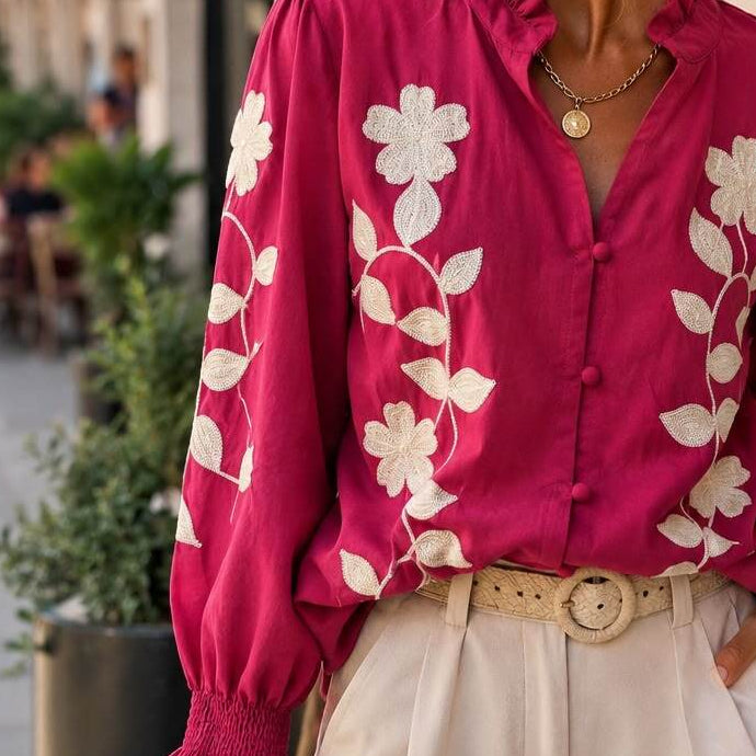 A woman wearing a vibrant magenta long-sleeve blouse with white floral embroidery, paired with cream-colored trousers and sunglasses, standing outdoors against a beige stone wall.