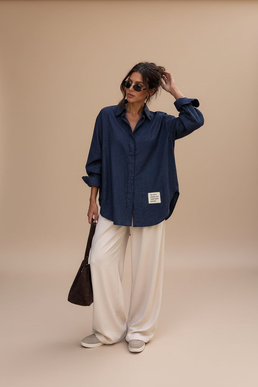A woman stands on a city sidewalk wearing an oversized navy blue pinstriped button-up shirt, cream wide-leg trousers, and beige slip-on shoes, while holding a brown shoulder bag.