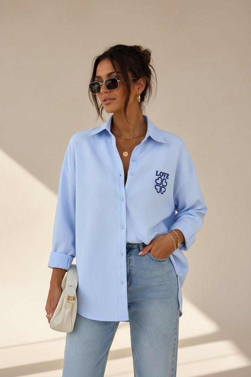 A woman wearing a light blue button-up shirt with a dark blue 'LOVE' graphic on the left chest, paired with light-wash jeans and holding a white clutch bag.