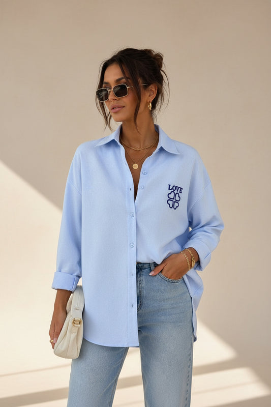 A woman wearing a light blue button-up shirt with a dark blue 'LOVE' graphic on the left chest, paired with light-wash jeans and holding a white clutch bag.