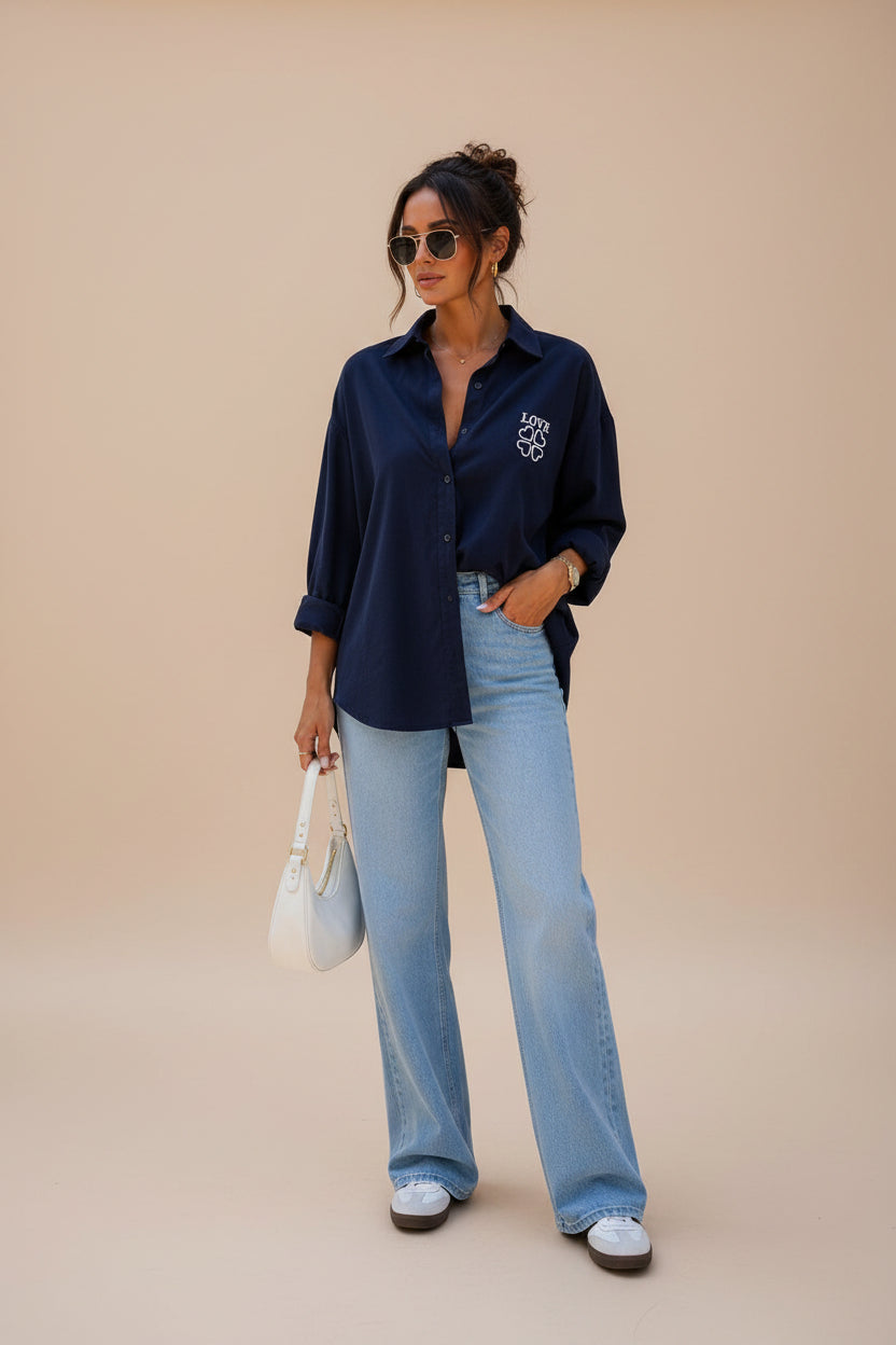 A woman stands on a city sidewalk wearing a navy blue button-up shirt with a white embroidered logo on the chest, light-wash wide-leg jeans, white sneakers, and holding a white handbag.