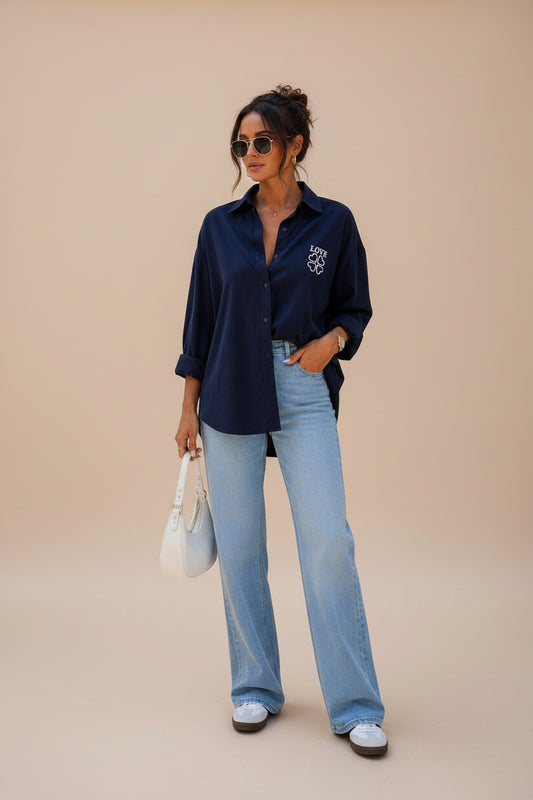 A woman stands on a city sidewalk wearing a navy blue button-up shirt with a white embroidered logo on the chest, light-wash wide-leg jeans, white sneakers, and holding a white handbag.