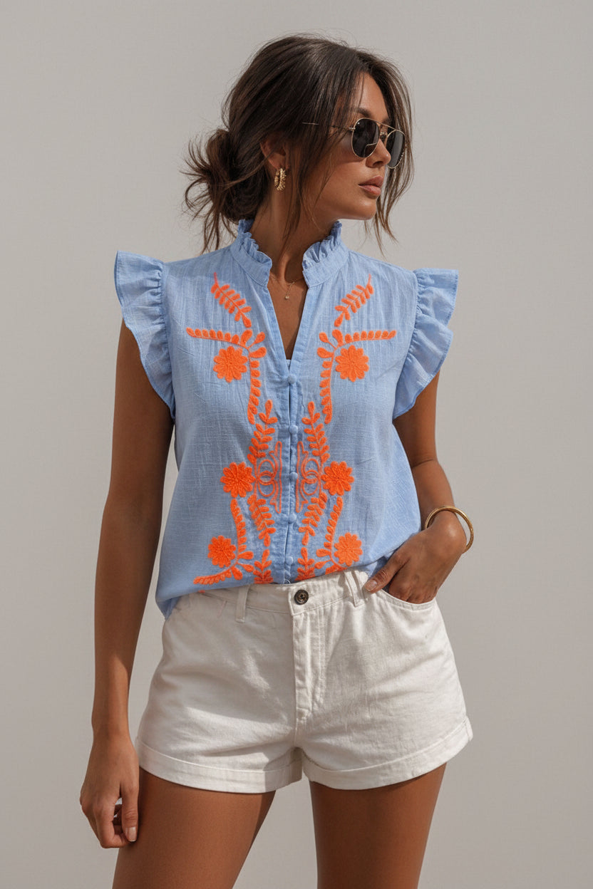 Woman wearing a light blue embroidered top with orange designs and white shorts against a neutral background.