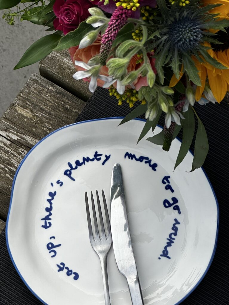 White plate with blue text, silver fork and knife, and colorful flowers on a wooden surface.