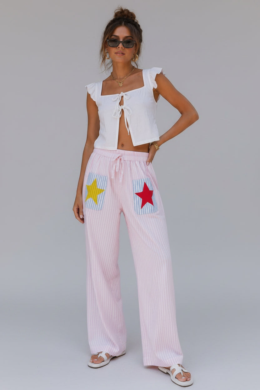 Woman wearing a white top and pink pants with star designs, standing next to a large potted plant.