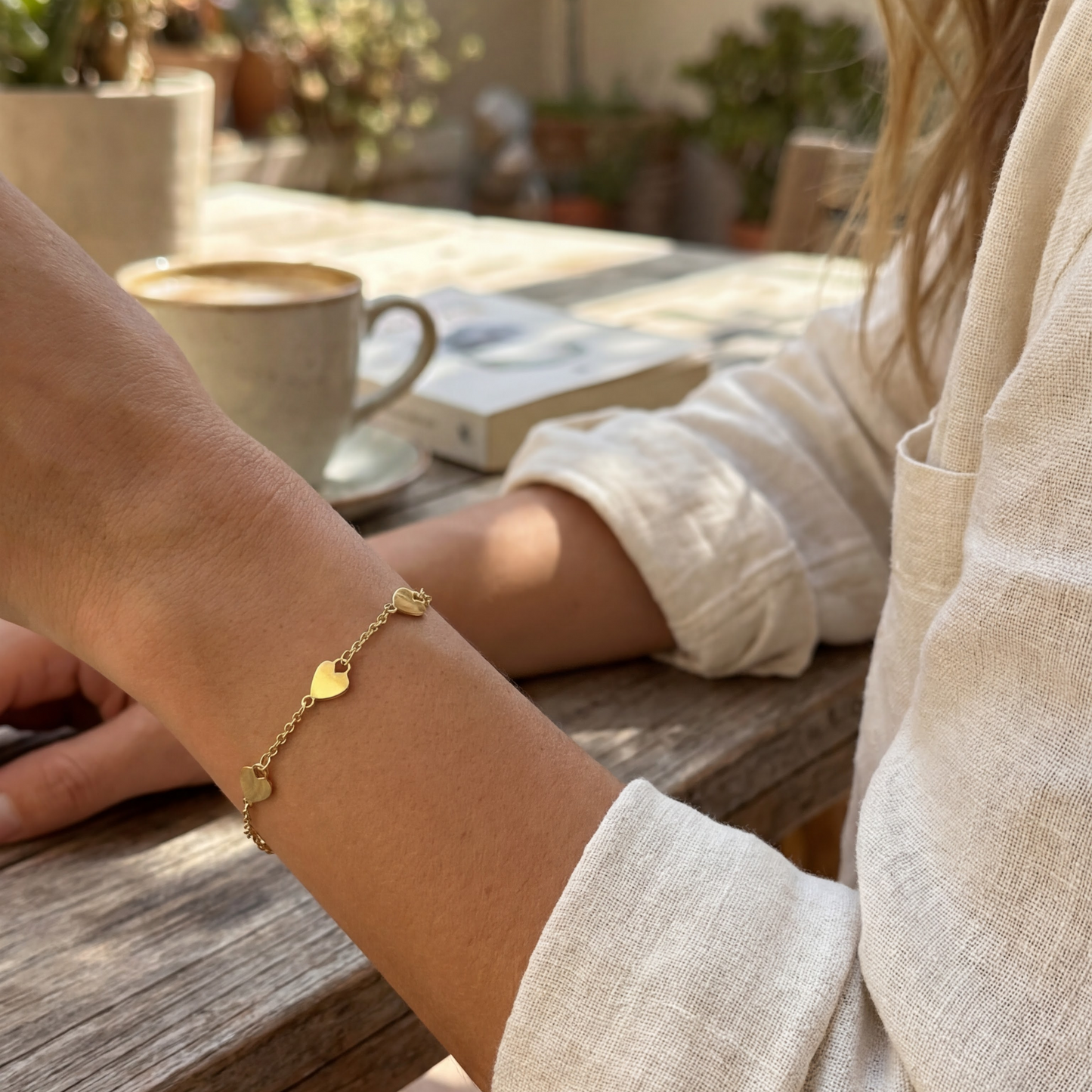 Person wearing a gold bracelet sitting at an outdoor table with a cup of coffee.